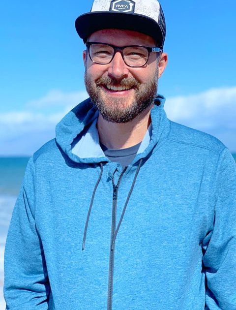 Jason Coffee in blue hoodie. Smiling at camera with sea in his background