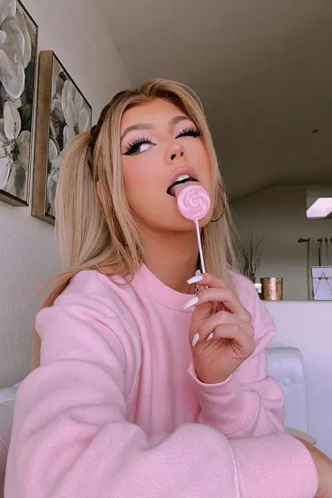 Loren Gray wearing pink top, winking, and licking a pink lollipop