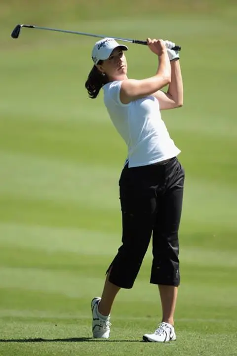 Paige Mackenzie playing golf and swinging her golf club. She is wearing white t shirt and black trousers