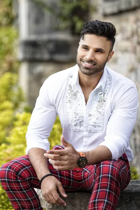 Rahim Pardesi is smiling and has perfect stubble. He is wearing embroidered white shirt and check red pants. His masculine arms can be seen.