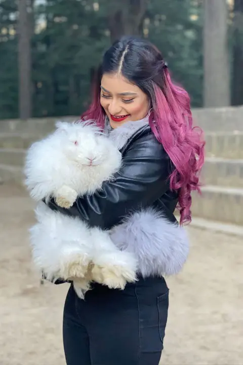 Anam Darbar holding her white pussy cat while looking at it and smiling happily. She is wearing tight black jeans and has pink stripe hair
