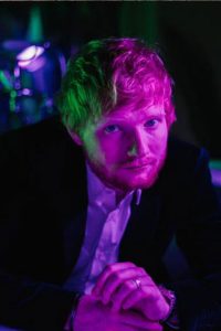 Ed Sheeran being hot and casual at party. He is looking intensely at camera with his green eyes